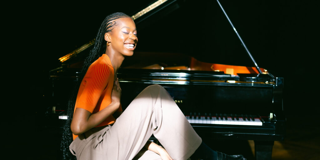 Black woman sits sideways at a piano and smiles broadly with eyes closed.