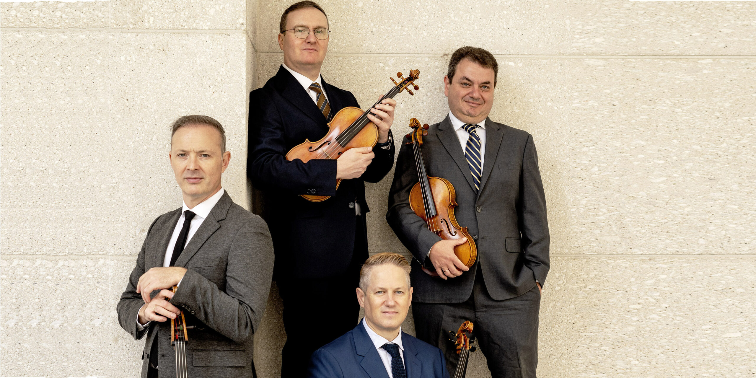 Four white men posing in front of an off-white stone wall. They are holding string instruments.