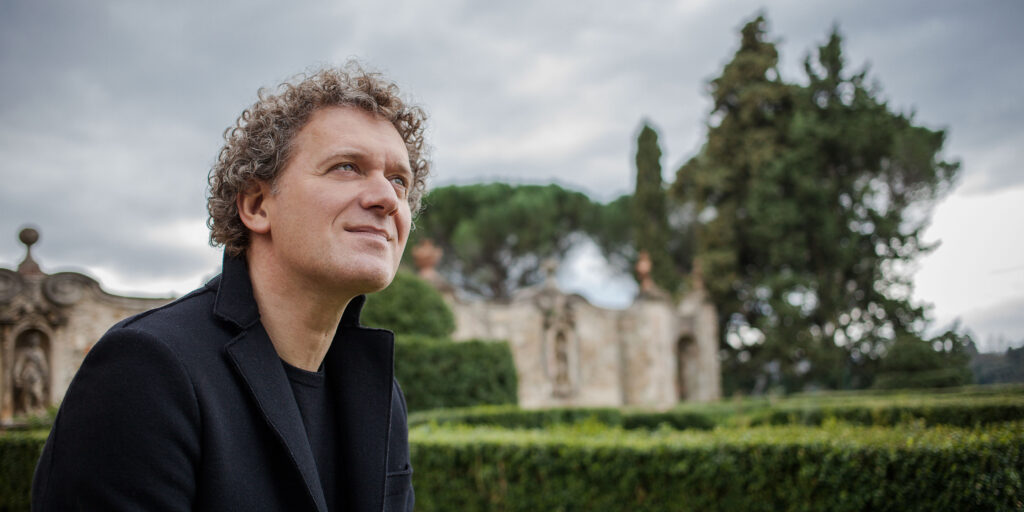 White man with short curly hair looks off into the distance. In the background is a manicured garden.
