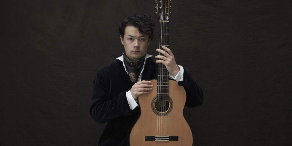 British-Japanese man with a serious expression posing with a guitar.