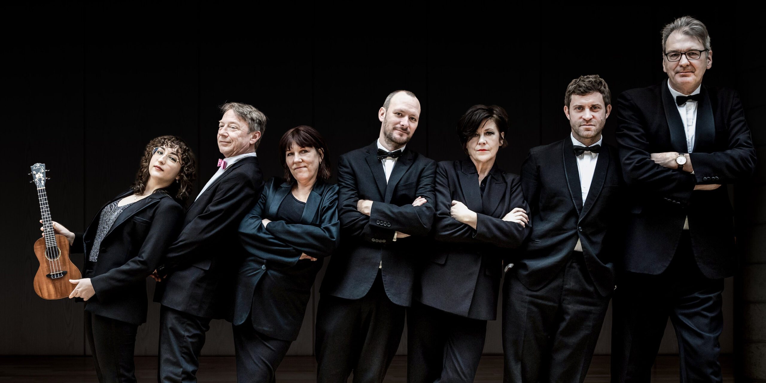 Group of men and women posing in suits. The person on the far left is holding a ukulele.