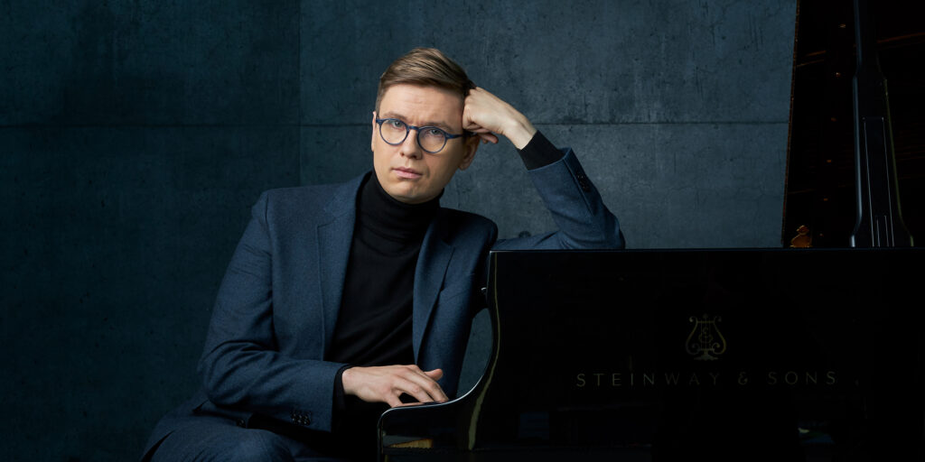 Man sits sideways and leans his elbow on the piano while looking at the camera.